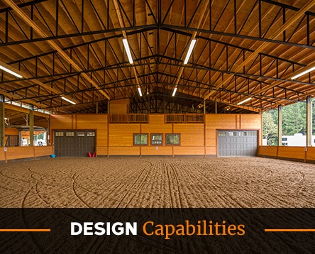 The airy interior of a spacious arena with wooden walls and ceiling features skylights and large garage-style doors. The sandy floor hints at equestrian activities, made possible by horse arena kits. Text overlay at the bottom reads "Design Capabilities.