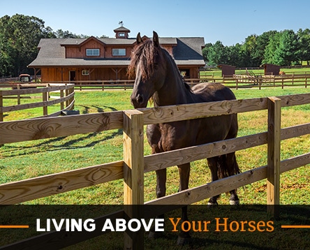 A dark brown horse stands in a wooden fenced pasture with a large barn, possibly built from an apartment barn kit, and trees in the background. The text at the bottom reads, "Living Above Your Horses.