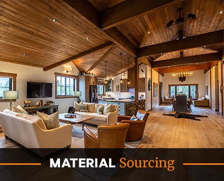 Spacious rustic living room with wooden ceiling and floor, featuring cream and brown armchairs, a TV, and a dining area. Large windows bring in natural light, enhancing the cozy charm of cabin kits. Text overlay says "Material Sourcing.