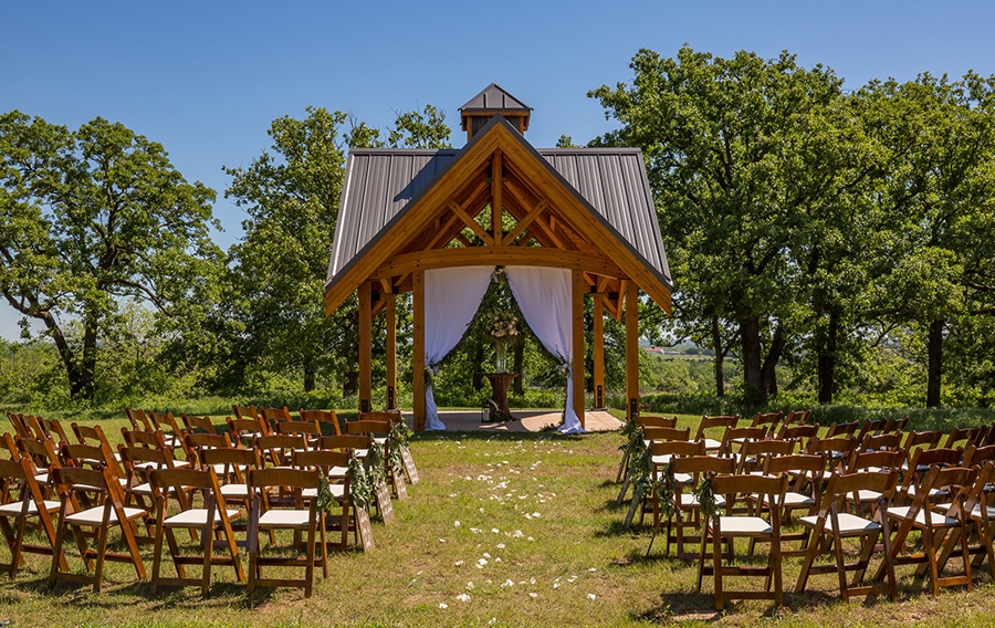 A rustic outdoor wedding setup features a wooden gazebo crafted from pavilion kits, elegantly draped with white fabric. Wooden chairs are arranged in rows on the grassy area, leading up to the gazebo. Lush green trees surround the scene under a clear blue sky.