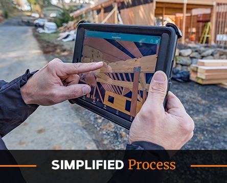 A person holds a tablet displaying construction plans for pavilion kits at a building site. The screen shows wooden beams within a structure. Wooden materials and a partially constructed building are visible in the background. The text "Simplified Process" is at the bottom.