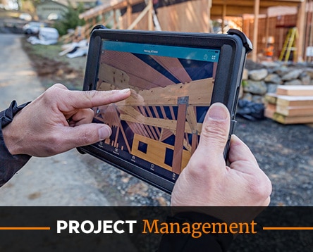 A person holds a tablet displaying architectural designs for barn home kits at a construction site. Wooden beams and construction materials are visible in the background. Text at the bottom reads "Project Management.