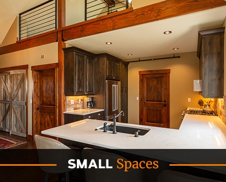 Modern kitchen interior with dark wood cabinets, white countertops, and a centrally located island with a sink. A barndoor and wood accents add rustic charm reminiscent of cozy cabin kits. Text overlay reads "SMALL Spaces" at the bottom.