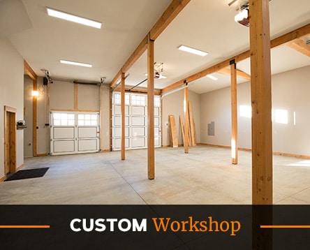 Spacious custom workshop with wooden beams, large garage doors, and bright overhead lights. The concrete floor and high ceiling create an open and versatile workspace, reminiscent of modern apartment barn kits. Text overlay reads "Custom Workshop" in white and orange.