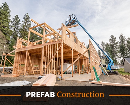 A multi-story wooden structure is under construction amidst a forest setting, reminiscent of horse arena kits. A worker on a blue lift is installing beams on the top level. The site is organized with construction materials and tools visible on the ground, reflecting precision craftsmanship.