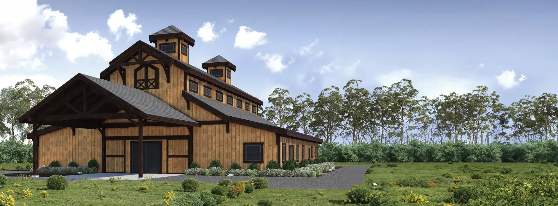 A large wooden barn with two cupolas, surrounded by green grass, flowering plants, and trees under a blue sky with scattered clouds.
