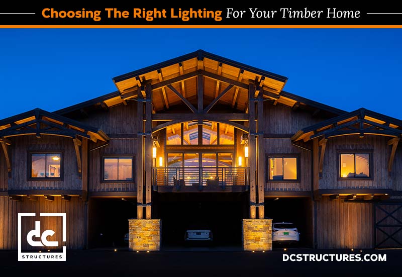 A timber home illuminated warmly at night. The house features large wooden beams, a gabled roof, and glowing interior lights visible through windows. Exterior lights highlight stone pillars. Text reads "Choosing the Right Lighting for Your Timber Home.