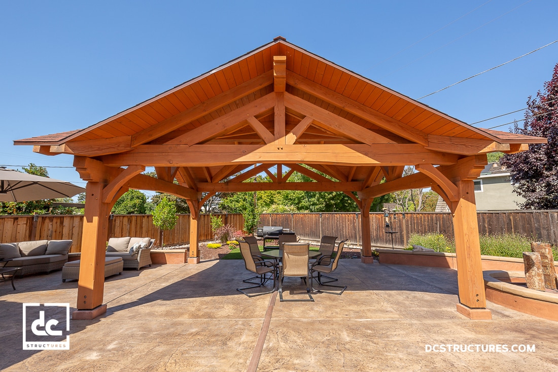 A wooden gazebo with a peaked roof covers a patio area with a table and chairs, surrounded by seating, plants, and a fence.