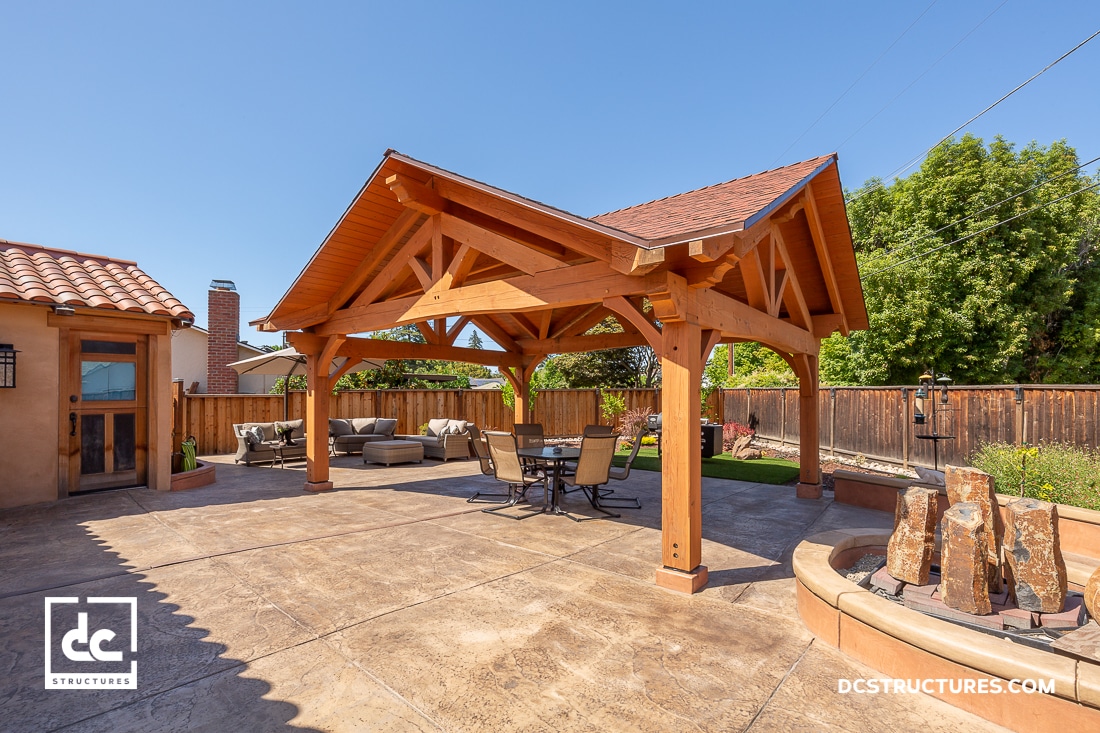 A wooden outdoor pavilion with exposed beams stands on a stamped concrete patio with patio furniture. A stone fountain is in front.