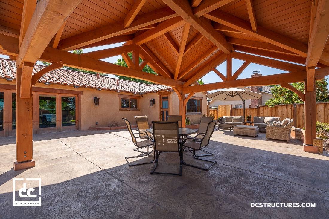 A spacious backyard patio with a wooden pavilion, central table and chairs, side seating area with umbrella, fenced by wood.