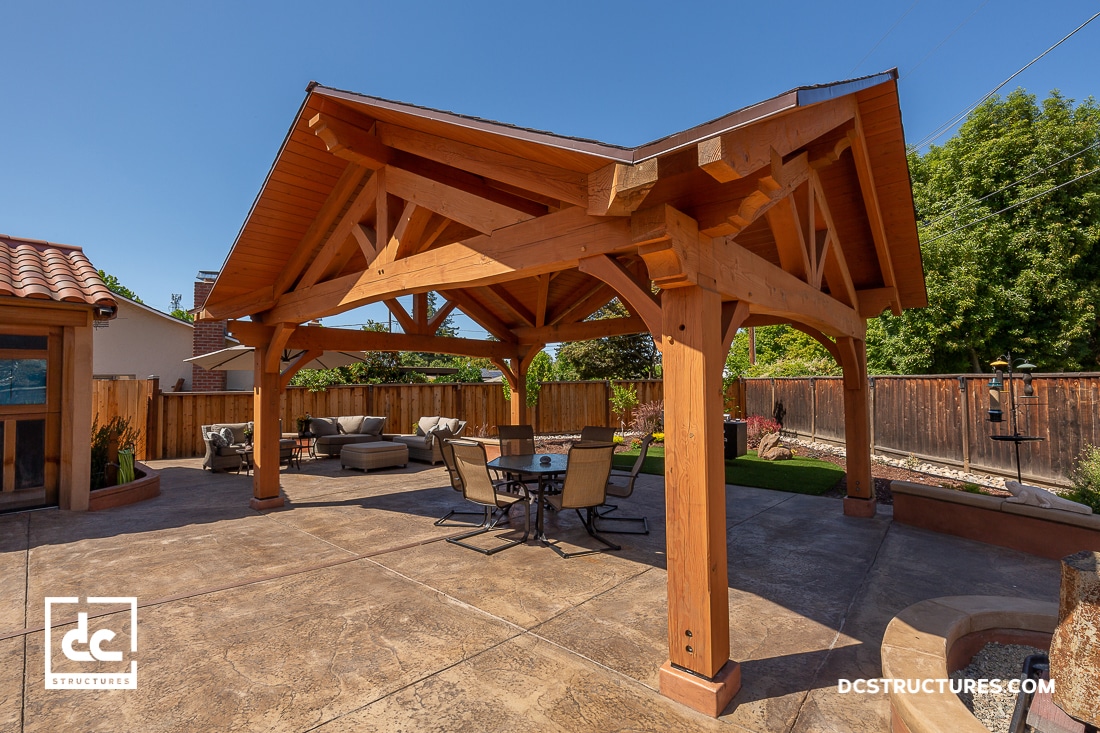 A spacious backyard has a wooden pavilion with exposed beams over a patio, with dining furniture and outdoor seating, trees, and a wooden fence.