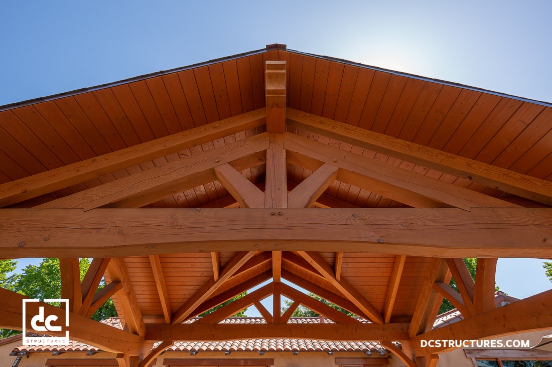 A close-up view of a wooden pergola roof with exposed beams, photographed from below against a clear blue sky.