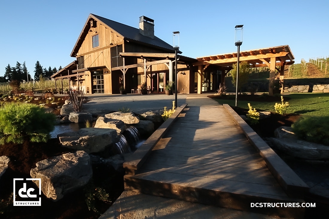 A rustic wooden barn-style building with a stone chimney, large windows, and covered patio, viewed from a bridge over a stream.