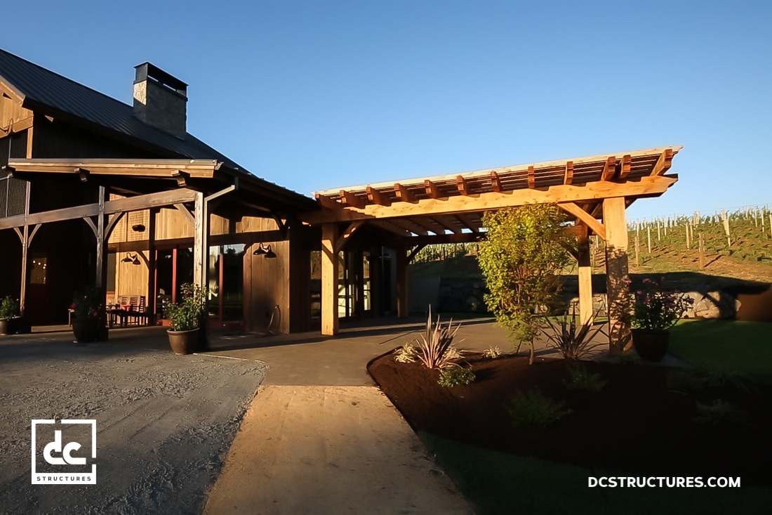 A modern barn-style building with wooden beams, large covered patio, landscaping, and vineyard in the background in warm sunlight.