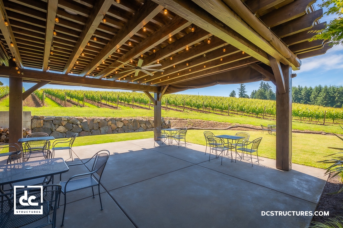 A covered patio with string lights and metal tables overlooks a scenic vineyard. A stone wall separates the patio from vineyard rows.