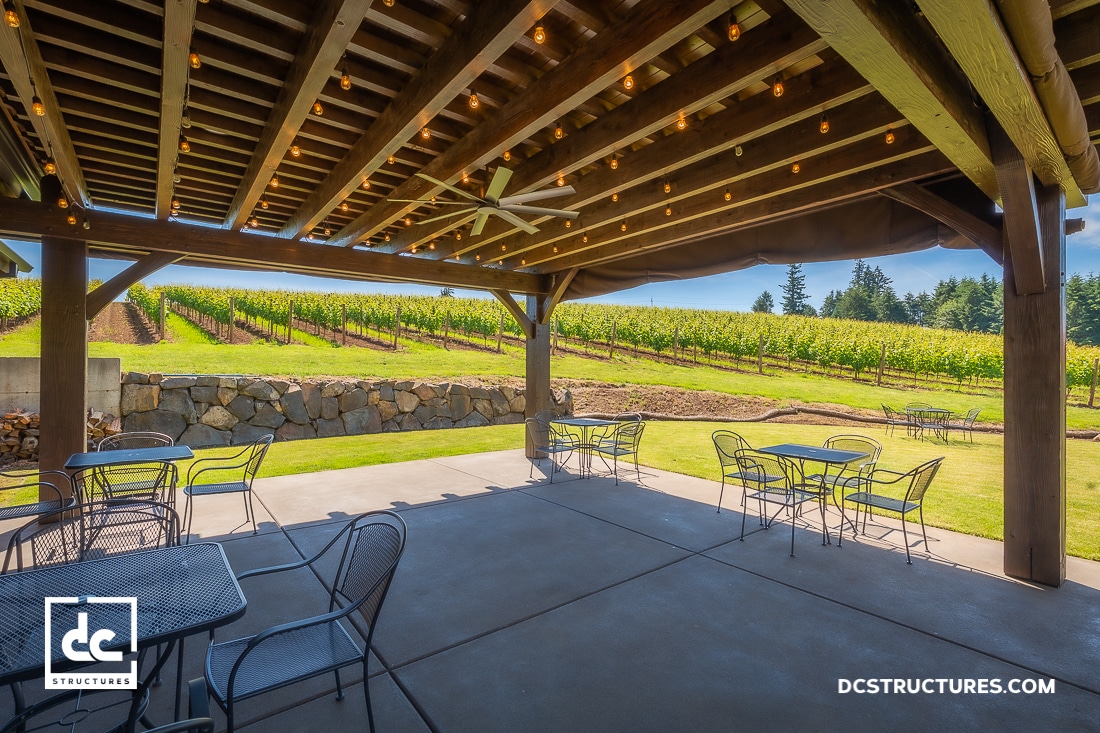 Covered patio with string lights, metal tables, and chairs overlooks a scenic vineyard, stone wall, grass, trees, and blue sky.