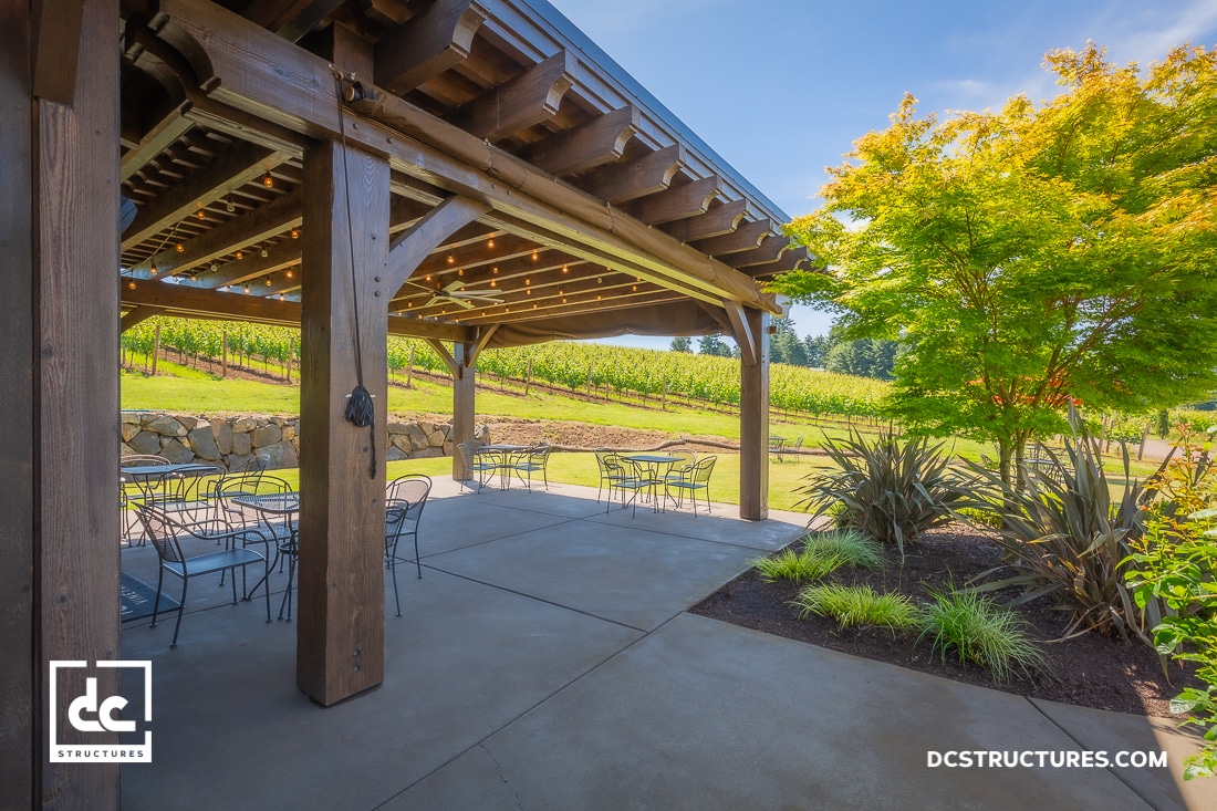 A wooden pergola with outdoor seating overlooks a vineyard on a sunny day. Lush green plants and a small tree border the patio.