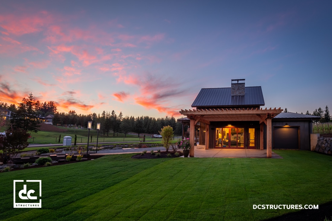 A small modern house with large windows and a wooden pergola sits on a manicured lawn at sunset, glowing warmly.
