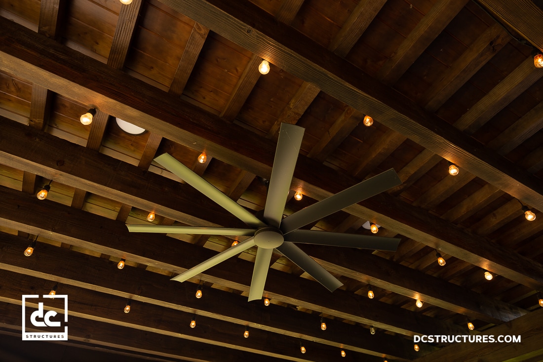 A large ceiling fan with multiple blades is mounted on a rustic wooden beam ceiling, surrounded by strings of small warm lights.