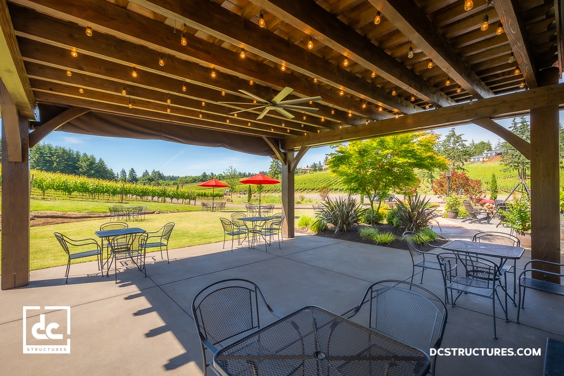 Covered patio with string lights, metal tables and chairs overlooking a vineyard with lush greenery, red umbrellas, and clear blue sky.
