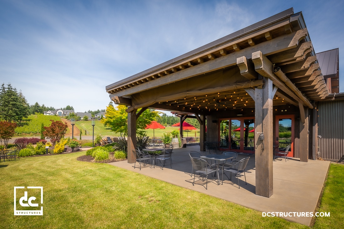 A covered outdoor patio with wooden beams, string lights, and metal tables and chairs overlooks a landscaped lawn and rolling hills.