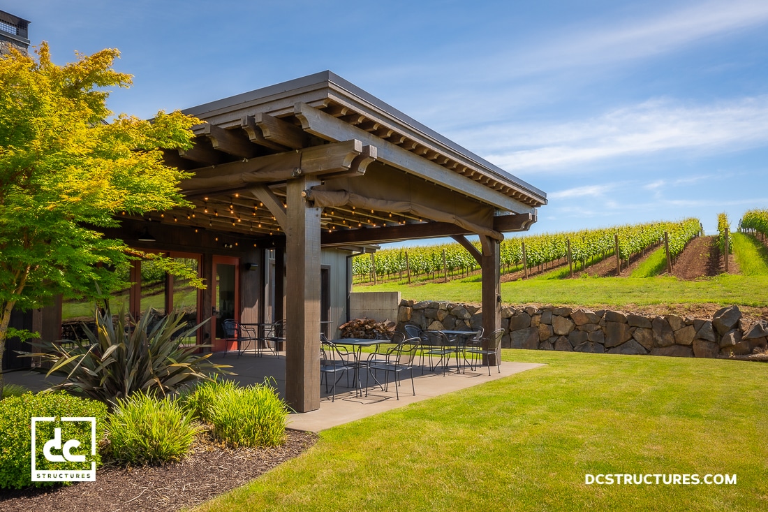 A modern wooden pergola with string lights shades an outdoor patio with tables and chairs, set beside a vineyard and stone wall under a blue sky.