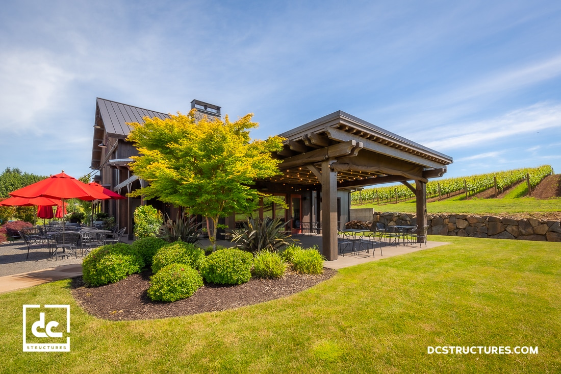 A modern barn-style building with a covered patio, surrounded by green grass, bushes, and a tree. Red umbrellas shade outdoor seating.