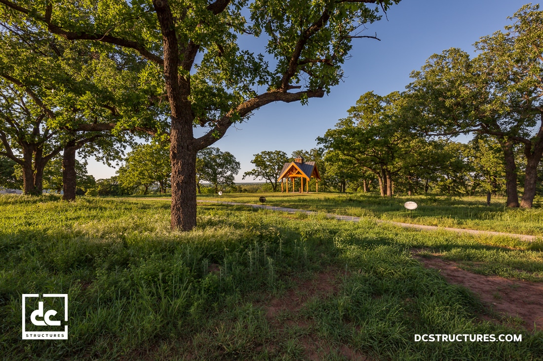 A small wooden pavilion with a peaked roof stands among tall green grass and large trees under a clear blue sky. A dirt pathway leads to the structure.