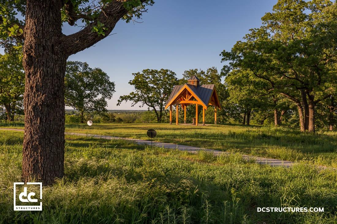 A small wooden pavilion with a metal roof stands in a grassy clearing surrounded by green trees under a blue sky. A concrete path leads to the pavilion.