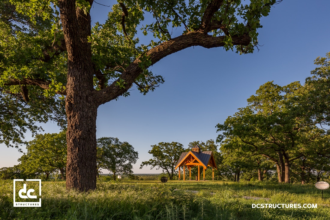 A small wooden pavilion with a dark roof stands among tall green trees and grass under a clear blue sky. Sunlight highlights the structure.