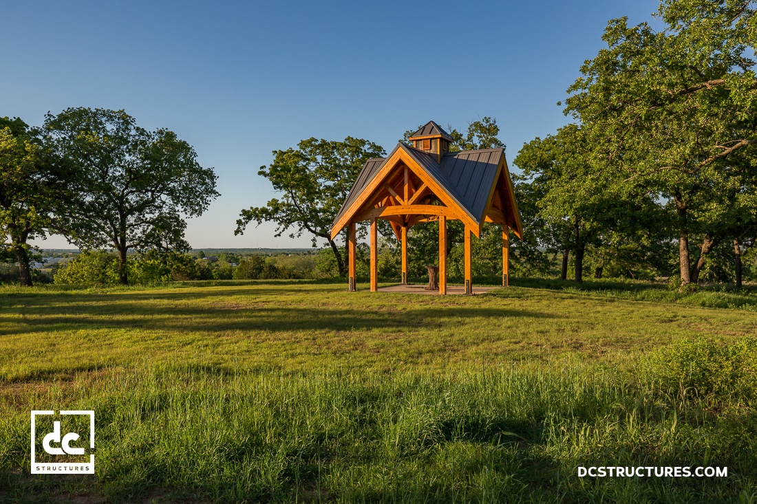 A small open wooden pavilion with a peaked roof stands in a grassy clearing surrounded by trees under a clear blue sky.