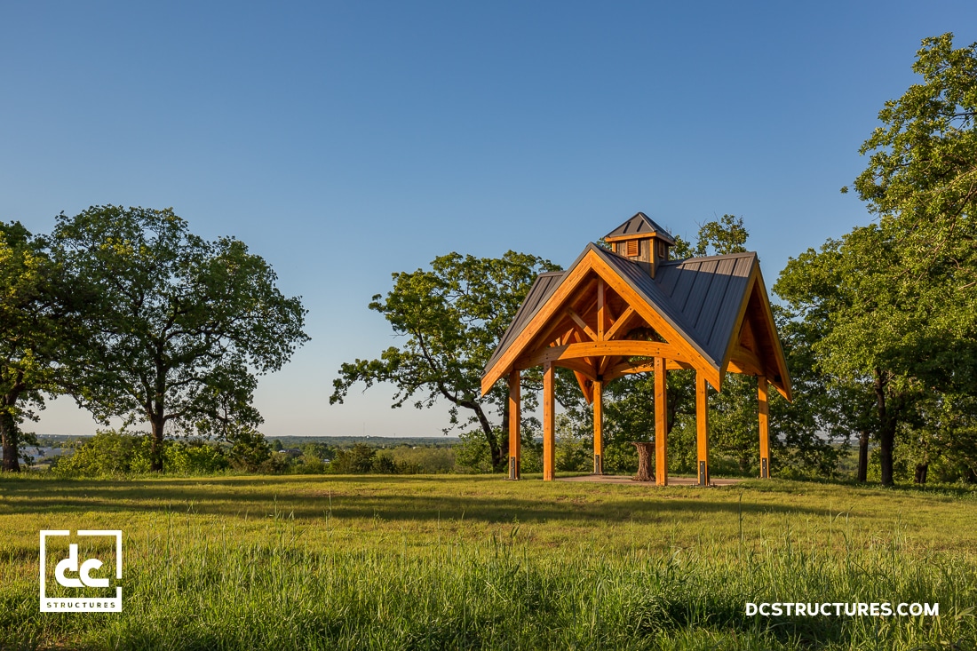A wooden pavilion with a steep metal roof stands on a grassy field surrounded by green trees under a clear blue sky.