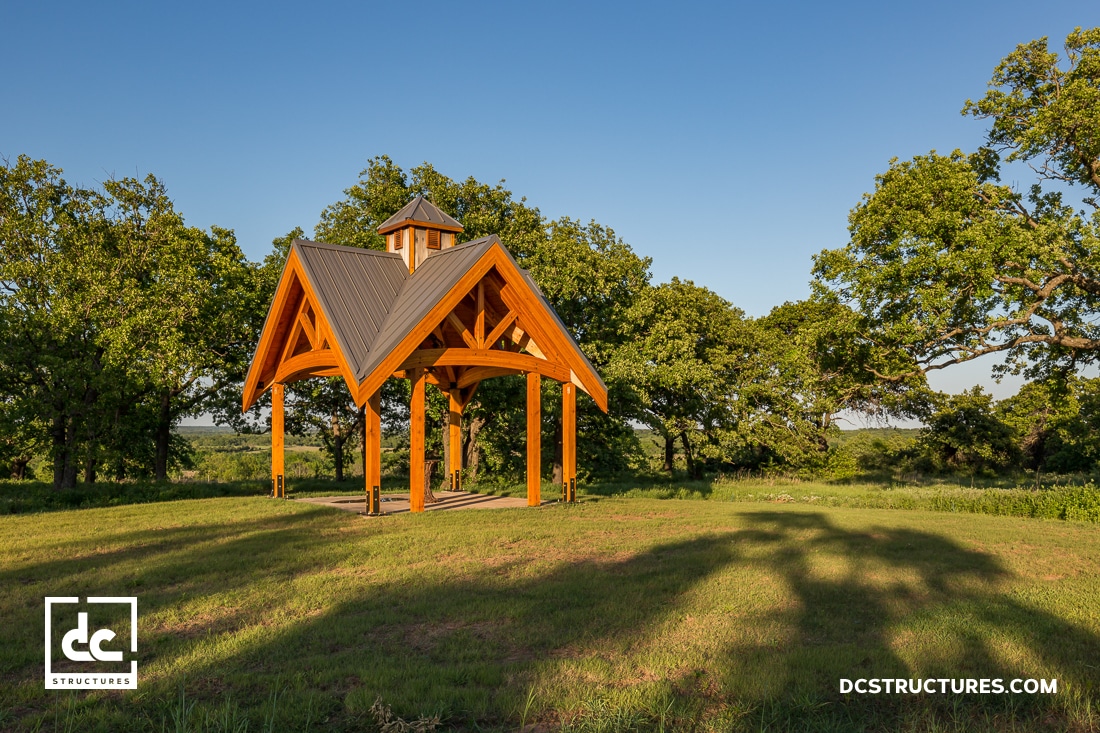 A wooden pavilion with a peaked metal roof stands on a grassy field surrounded by trees under a clear blue sky.