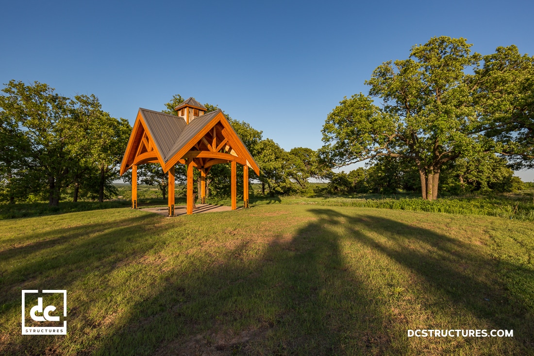 A wooden pavilion with a pitched roof stands on a grassy field surrounded by green trees under a clear blue sky. Shadows stretch across the grass.