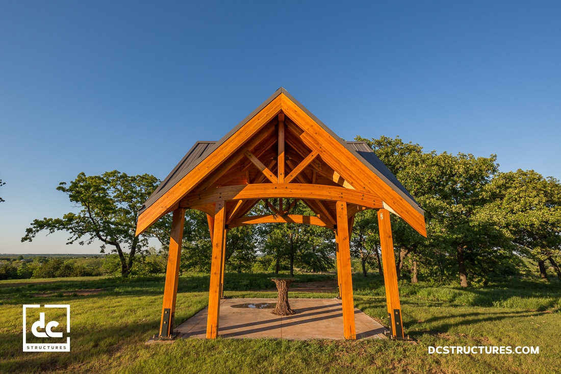 A wooden pavilion with a peaked roof and open sides sits on a concrete pad in a grassy field, surrounded by trees and blue sky.