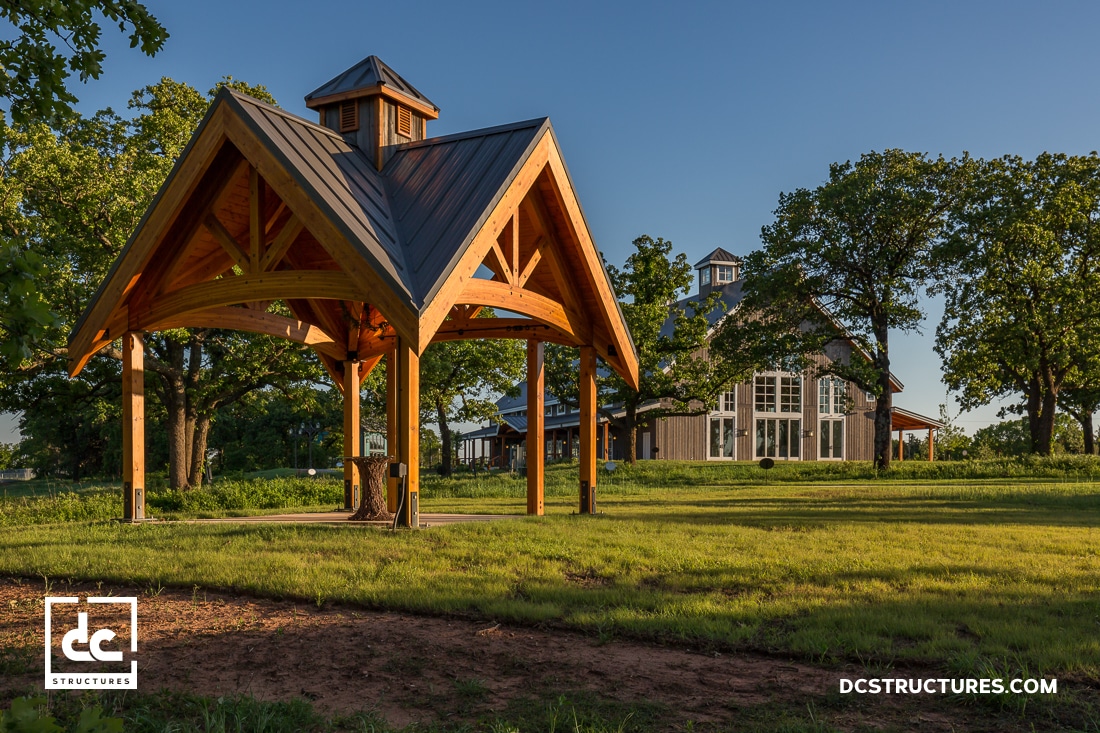 A wooden pavilion with a metal roof stands on a grassy lawn near a modern barn-style building surrounded by trees in late afternoon light.