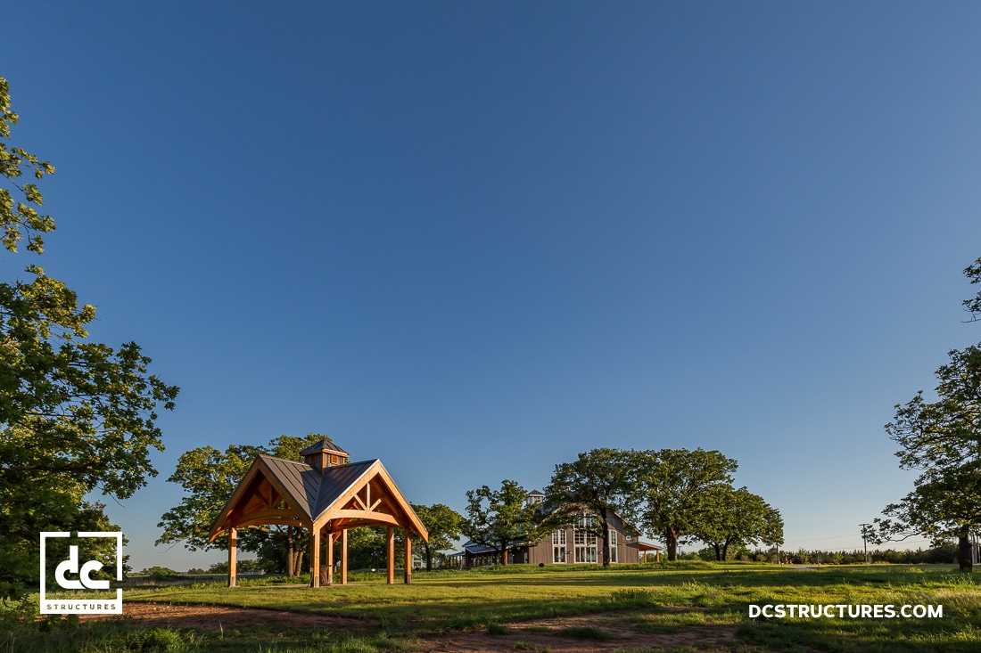 A wooden open-air pavilion stands on grass under a clear blue sky, with a larger barn-style building and trees in the background.