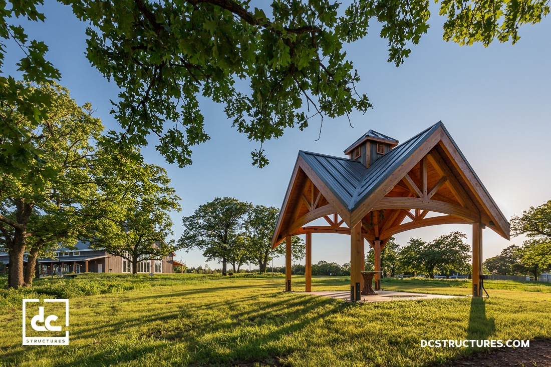 A wooden pavilion with a metal roof stands on a grassy lawn, surrounded by trees under a clear sky. Sunlight casts long shadows.