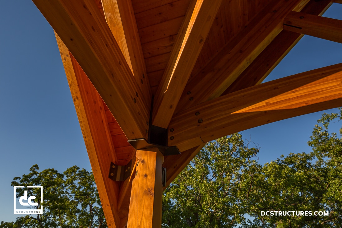 Close-up view of a wooden timber frame structure with metal brackets, set against a clear blue sky and trees in the background.