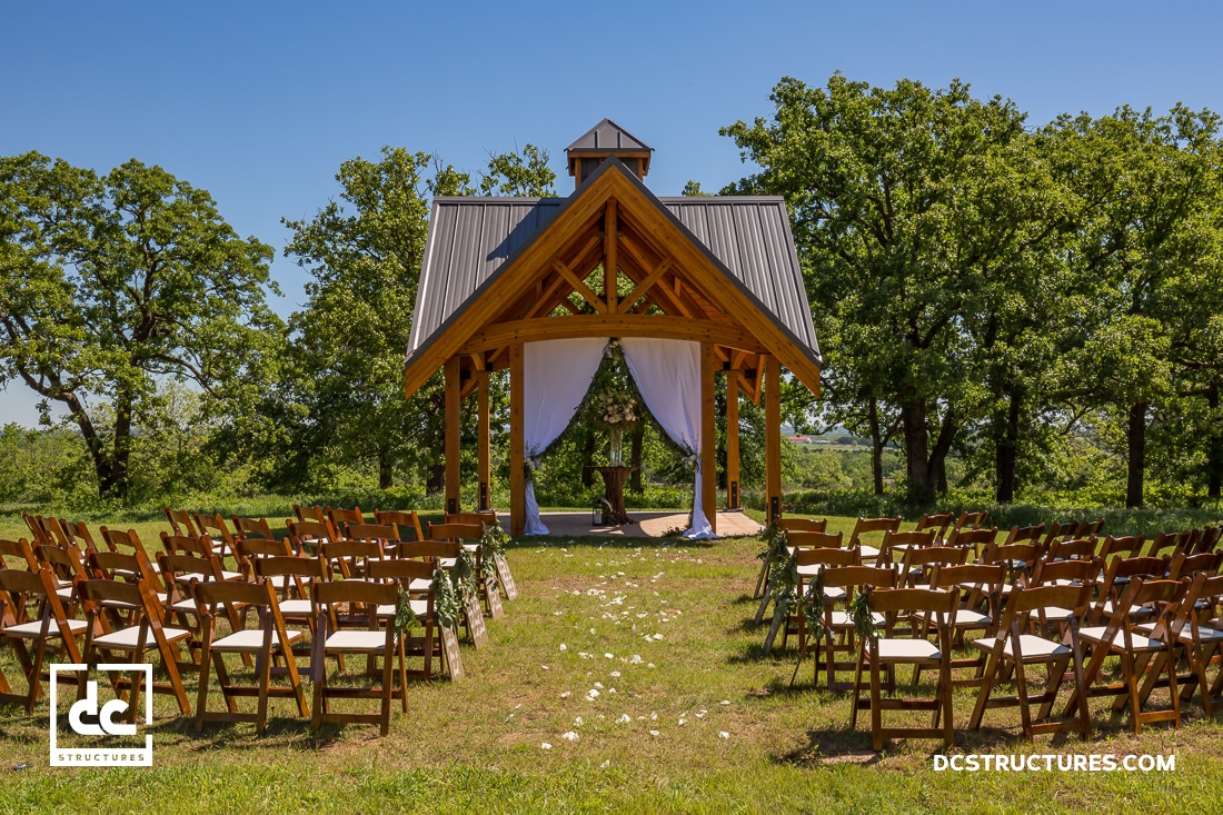 An outdoor wedding venue with wooden chairs facing a pavilion draped with white curtains, surrounded by grass and trees.