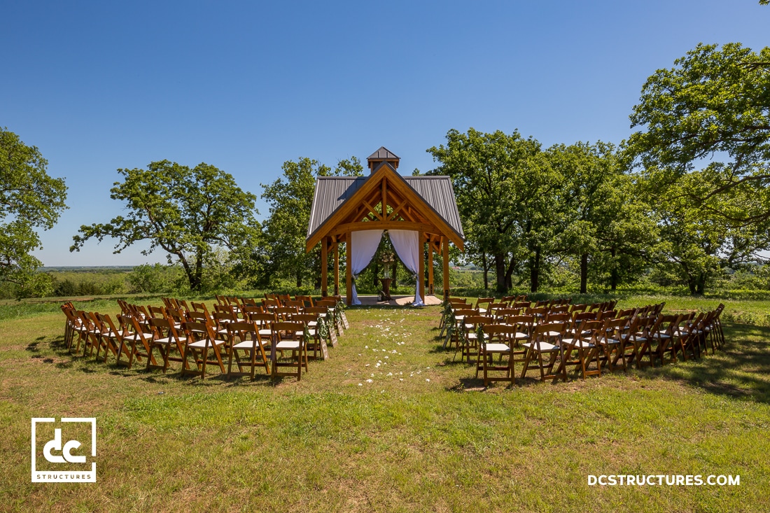 Outdoor wedding ceremony setup with rows of wooden chairs facing a small open wooden pavilion, surrounded by grass and trees under a clear blue sky.