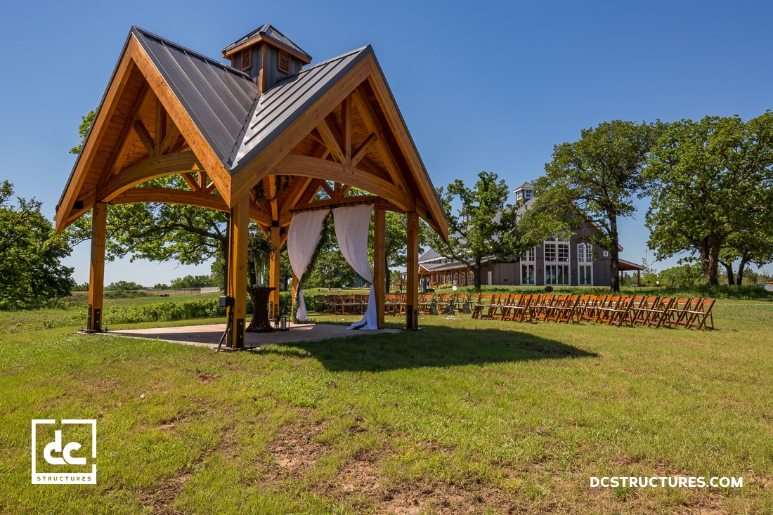 A wooden pavilion with a peaked roof stands on a grassy lawn, draped with white curtains. Rows of chairs face the pavilion.