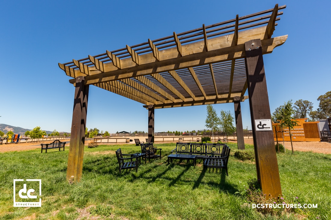 A wooden pergola stands on a grassy area with outdoor seating underneath, surrounded by benches, trees, and a clear blue sky.