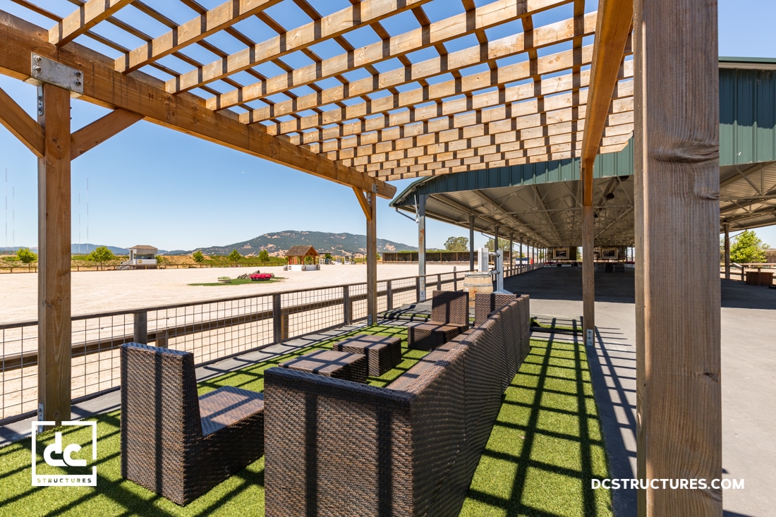 Outdoor seating area with wicker furniture under a wooden pergola, overlooking an open, sunny landscape with mountains in the background.