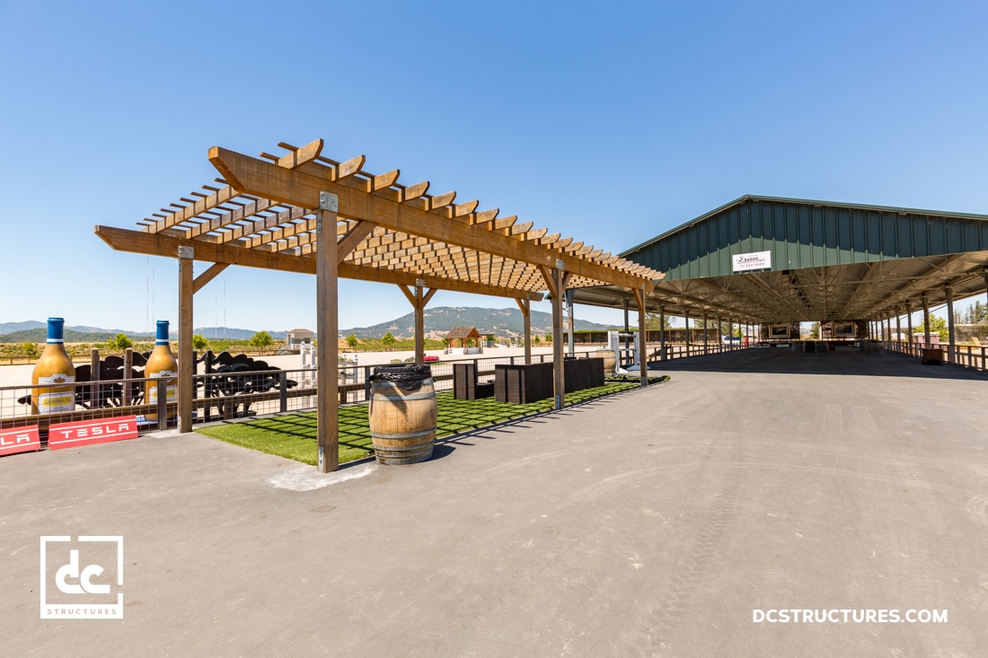 A wooden pergola with a picnic area and wine barrels stands beside a large open-sided barn under a clear blue sky. Mountains are visible in the background.