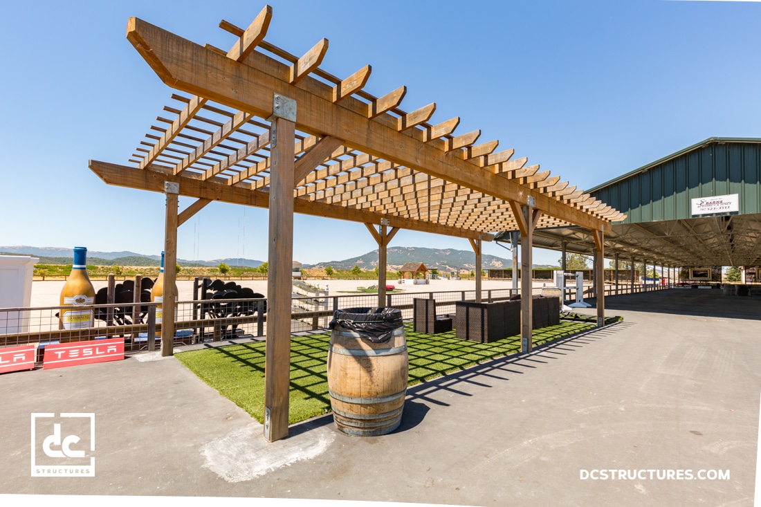 A wooden pergola with benches and a barrel trash bin stands on a paved area with artificial grass near mountains under a clear blue sky.