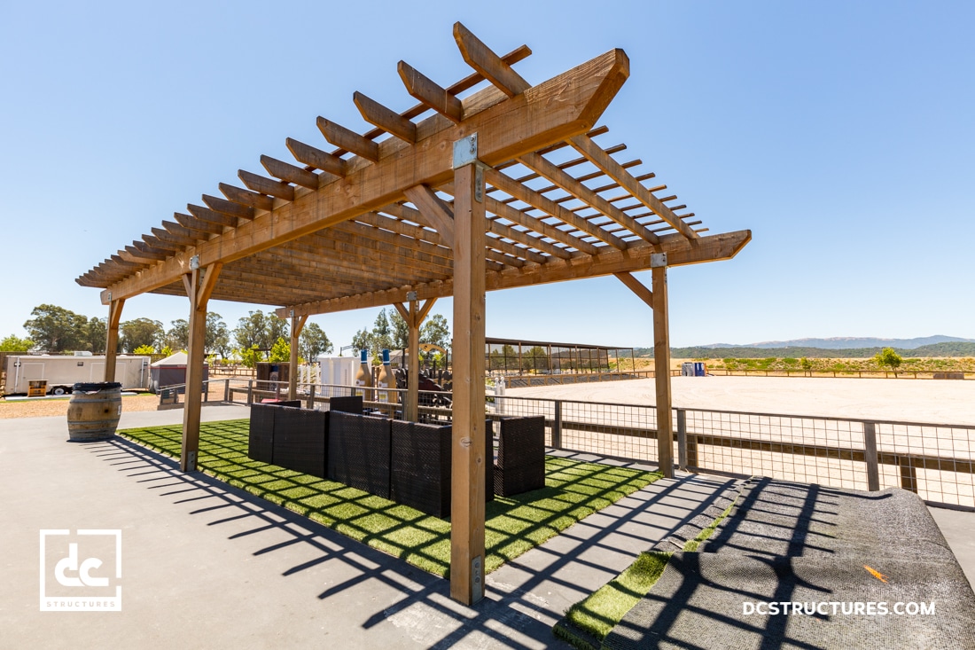A wooden pergola stands over outdoor seating on artificial grass, under a clear blue sky with distant hills. Barrels and patio furniture sit beneath the structure inside a fenced area.