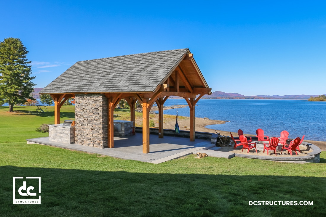 A wooden and stone pavilion with a barbecue area sits on a lawn by a lake. Nearby, red Adirondack chairs surround a fire pit under a clear blue sky.