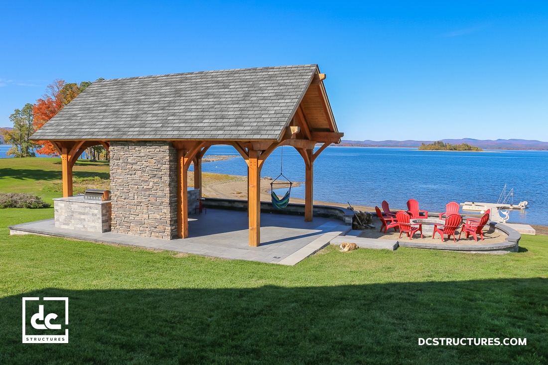 A lakeside pavilion with a wooden roof and stone wall stands on grass. Red Adirondack chairs circle a fire pit nearby.