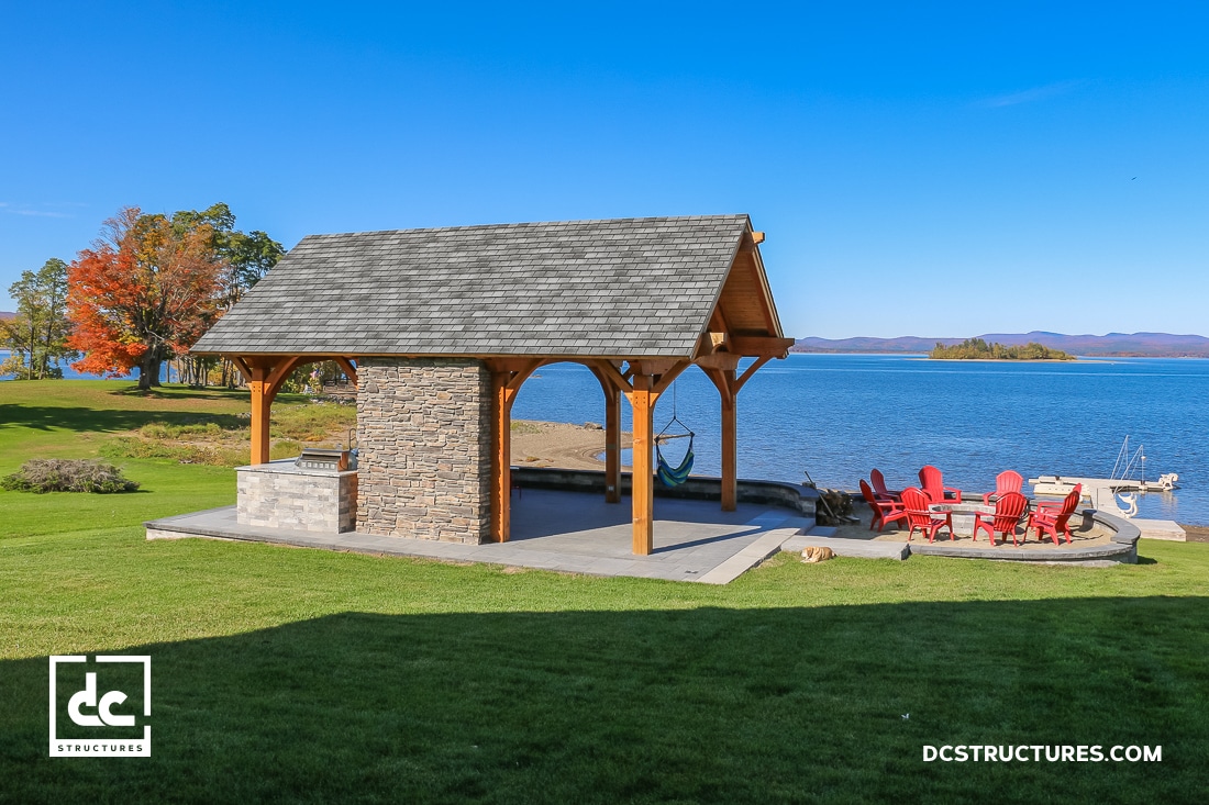 A lakeside pavilion of stone and wood sits on a grassy lawn. Red Adirondack chairs circle a firepit, with a hammock nearby.